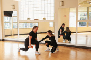 Client Working with Personal Trainer in a light filled studio doing a lunge and holding orange weights