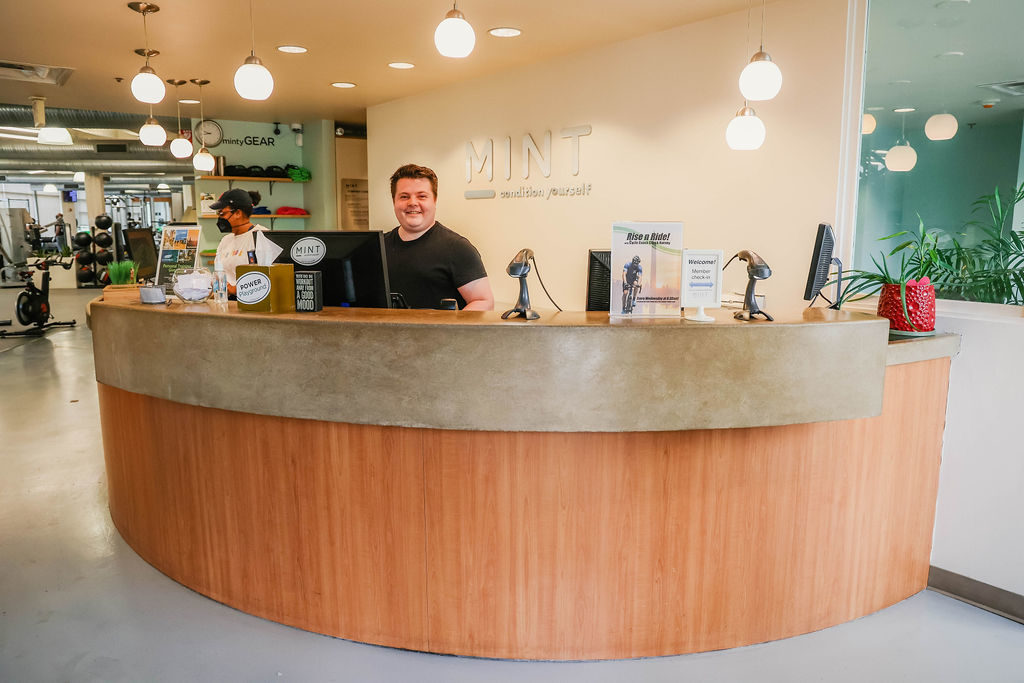 MINT Studio Assistant Standing in front of a computer at the front desk entrance smiling.